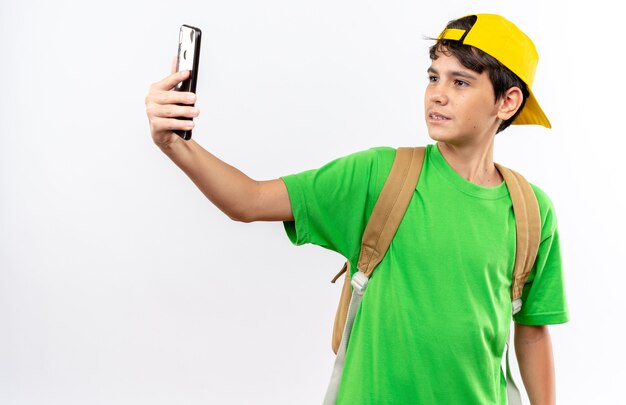 Niño de escuela joven satisfecho con mochila con gorra tomar un selfie aislado en la pared blanca