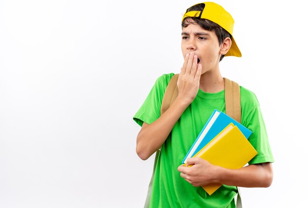 Niño de escuela joven sorprendido con mochila con gorra sosteniendo libros con la boca cubierta con la mano aislada en la pared blanca con espacio de copia