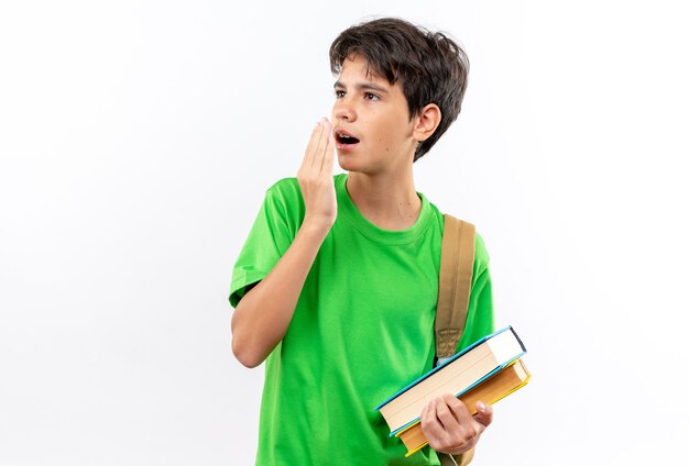 Niño de escuela joven sorprendido con mochila sosteniendo libros con la boca cubierta con la mano aislada en la pared blanca