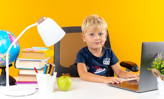 Niño de escuela pequeño disgustado sentado a la mesa con herramientas escolares utiliza portátil