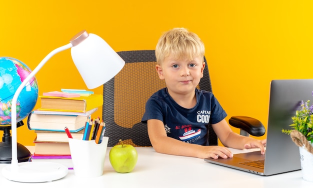 Niño de escuela pequeño disgustado sentado a la mesa con herramientas escolares utiliza portátil