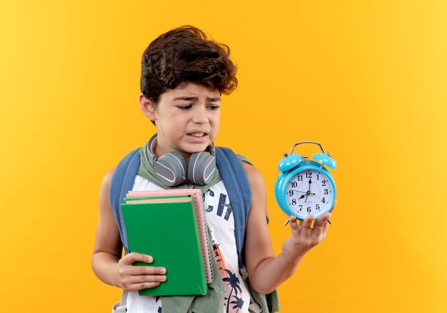 Niño de escuela preocupado con mochila y auriculares sosteniendo libros y mirando el despertador en su mano aislado sobre fondo amarillo