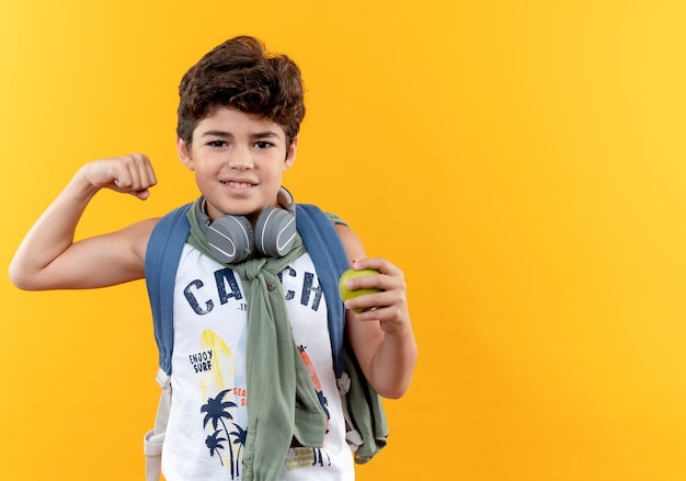 Niño de escuela satisfecho con bolsa trasera y auriculares sosteniendo apple y mostrando un gesto de sí aislado sobre fondo amarillo con espacio de copia