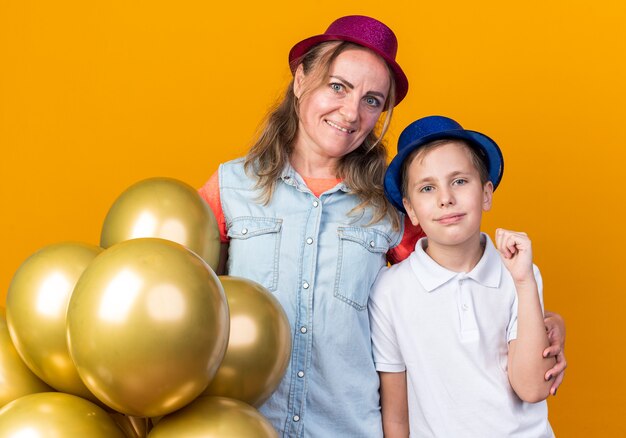 Niño eslavo joven complacido con sombrero de fiesta azul manteniendo el puño y de pie con su madre con sombrero de fiesta púrpura sosteniendo globos de helio aislados en la pared naranja con espacio de copia