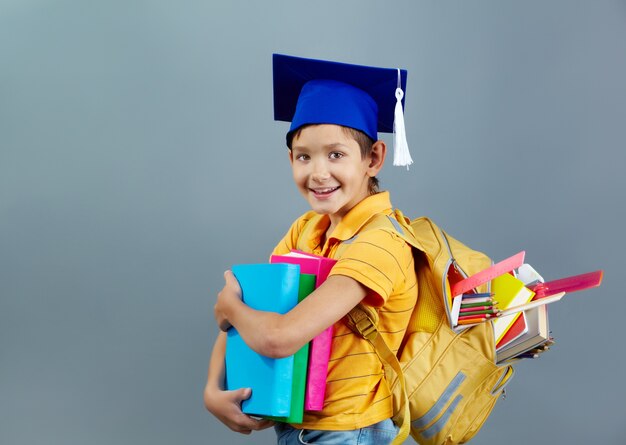 Niño exitoso con gorro de graduación y mochila llena de libros