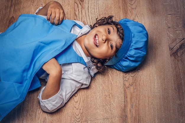 Foto gratuita un niño feliz con el pelo rizado castaño vestido con un uniforme de cocinero azul tirado en un suelo de madera.