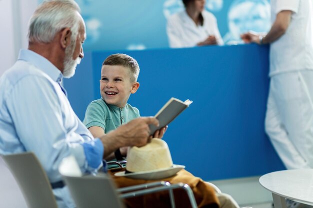 Un niño feliz y su abuelo leyendo un libro y comunicándose mientras esperan una cita médica en la clínica