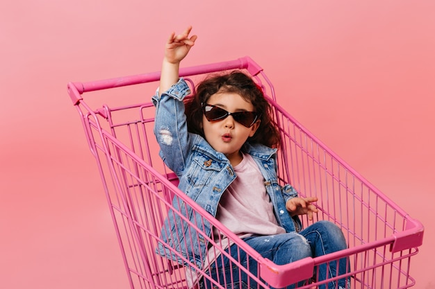 Niño gracioso con gafas de sol sentado en el carrito de compras. Disparo de estudio de niña feliz en chaqueta vaquera.