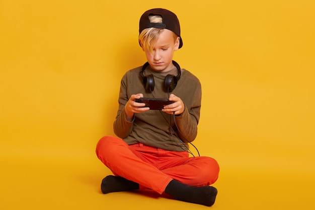 Niño jugando en el teléfono mientras está sentado en el piso aislado en amarillo, niño masculino con teléfono móvil en las manos, posando con auriculares alrededor del cuello, jugando juegos en línea.