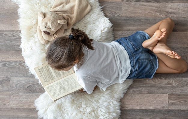 El niño lee un libro acostado en una acogedora alfombra en casa con su osito de peluche favorito.