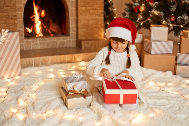 Niño lindo abriendo la caja presente de Santa Claus, vestido con suéter blanco y sombrero de santa claus, posando en la sala festiva con chimenea y árbol de Navidad mientras está sentado en un piso blando.