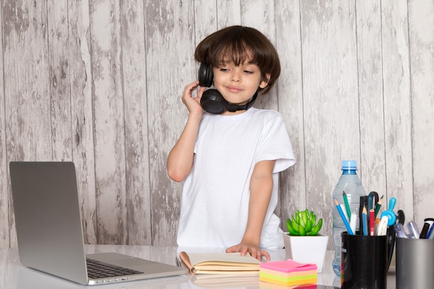 Foto gratuita niño lindo niño en camiseta blanca auriculares negros con portátil gris sobre la mesa junto con planta verde