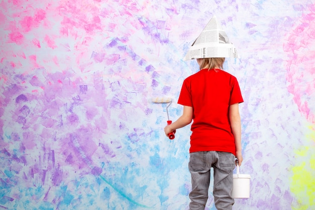 Foto gratuita niño lindo niño vista posterior en camiseta roja y jeans grises que pintan la pared colorida