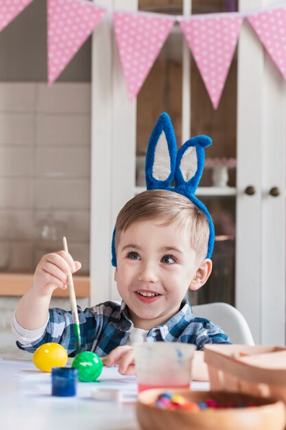 Niño lindo con orejas de conejo pintando huevos de pascua