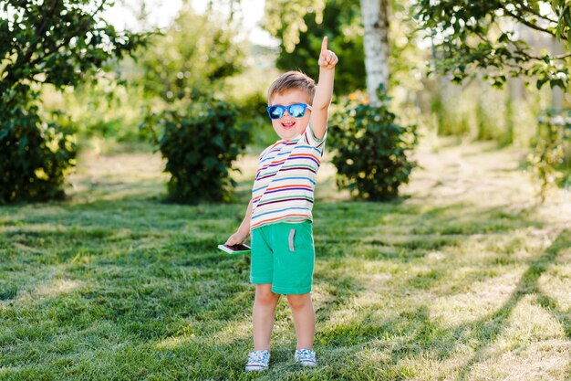 El niño lindo parece inspirado y feliz con gafas de sol brillantes con su teléfono.