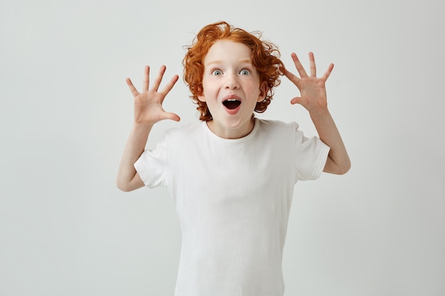 Niño lindo con el pelo de jengibre en camiseta blanca divirtiéndose en casa, haciendo estallar los ojos con la boca abierta