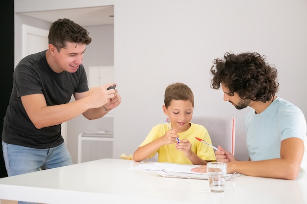 Niño lindo positivo haciendo tareas escolares en casa con la ayuda de dos papás, escribiendo en papeles. Hombre tomando fotografías de su familia. Concepto de familia y padres gay