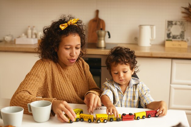 Niño lindo que disfruta del juego sentado con su madre alegre en la mesa de la cocina durante el desayuno. Retrato de familia de joven mujer latina jugando con su adorable hijo. Infancia, juegos e imaginación