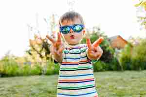 Foto gratuita niño lindo que se divierte y se ve muy serio con sus gafas de sol en el jardín.