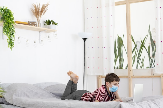 Niño con máscara médica viendo algo en la computadora portátil en la cama