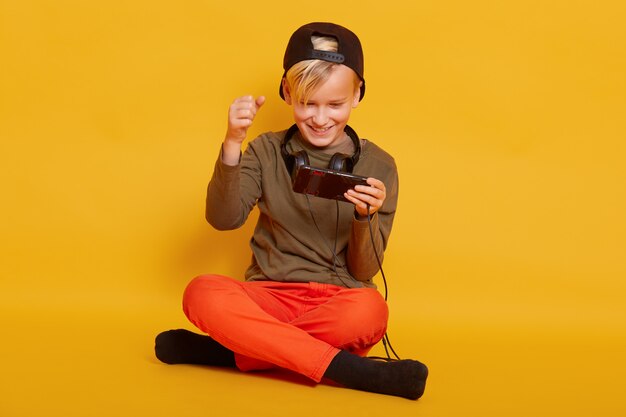 niño masculino jugando en el teléfono celular mientras está sentado en el piso aislado en amarillo, jugando su juego en línea favorito por teléfono, mantiene las piernas cruzadas, aprieta los puños.