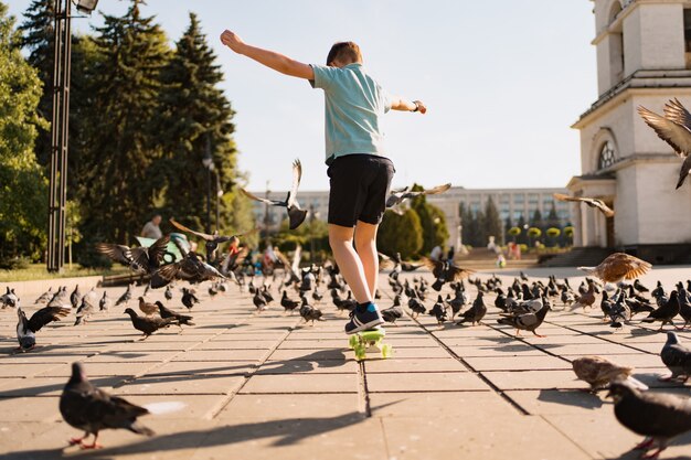 Un niño montando penny board en el parque en un cálido verano con palomas y cielo