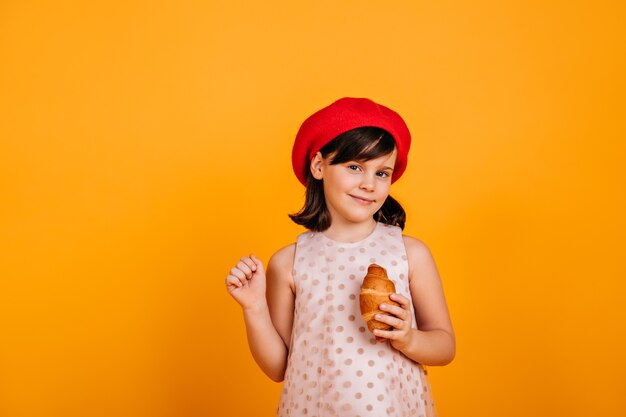 Niño morena curioso posando en la pared amarilla. niña preadolescente comiendo croissant.