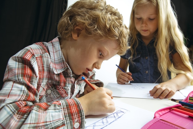 Niño y niña preparándose para la escuela después de un largo receso de verano. De vuelta a la escuela. Pequeños modelos caucásicos dibujando juntos sobre fondo blanco y negro. Concepto de infancia, educación, vacaciones o deberes.