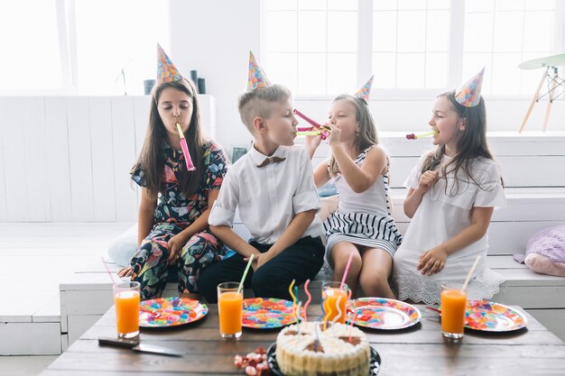 Niño y niñas soplando cuernos partido cerca de la torta