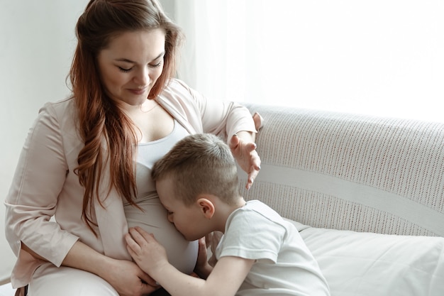 Niño niño abrazando y besando el vientre de su madre embarazada en el sofá de casa.