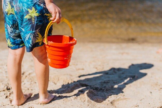 Niño en pantalones cortos con cubo de juguete en la playa del mar