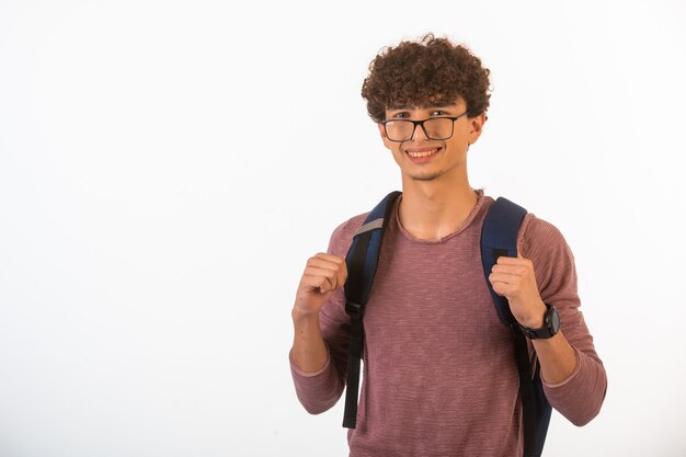 Niño de pelo rizado con gafas ópticas sosteniendo su mochila es feliz y alegre, vista frontal.