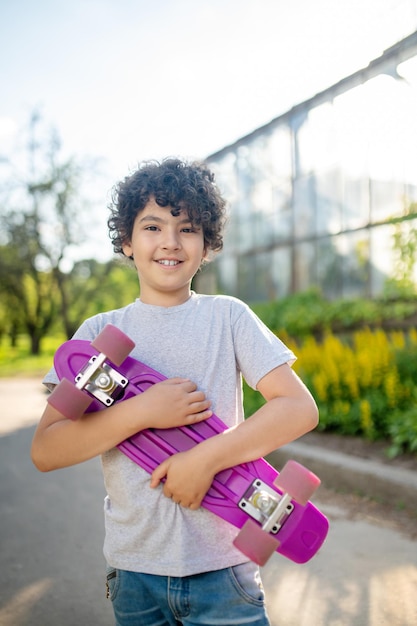 Niño de pelo rizado sonriente y complacido parado en la calle y sosteniendo su patineta con ambas manos