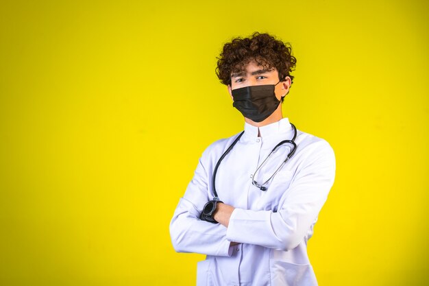 Niño de pelo rizado en uniforme médico blanco con estetoscopio con mascarilla