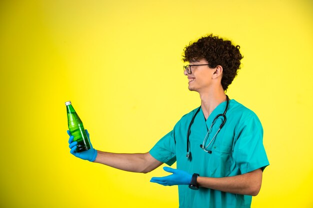 Niño de pelo rizado en uniforme médico y máscaras de mano mirando a una botella de líquido y sonriendo.