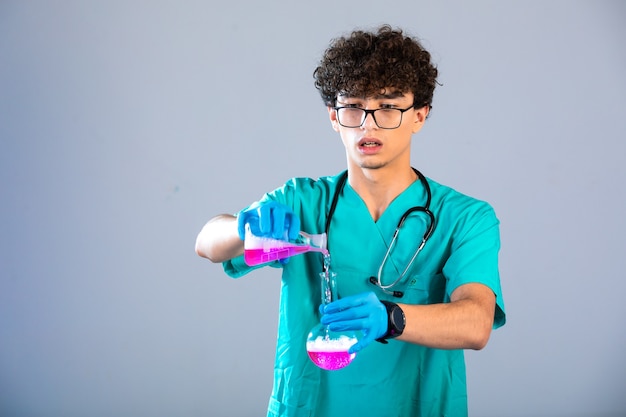 Niño de pelo rizado en uniforme médico y máscaras de mano poniendo líquido rosa de un matraz a otro