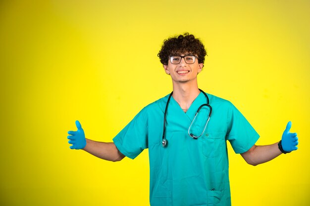 Niño de pelo rizado en uniforme médico y máscaras de mano con los pulgares para arriba.