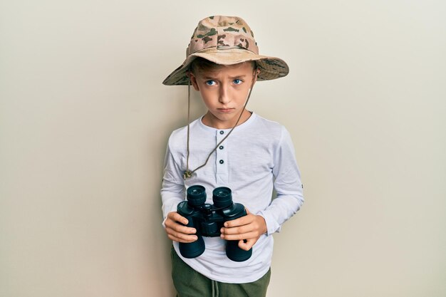 Niño pequeño caucásico con sombrero de explorador sosteniendo binoculares escéptico y nervioso, frunciendo el ceño molesto por el problema. persona negativa