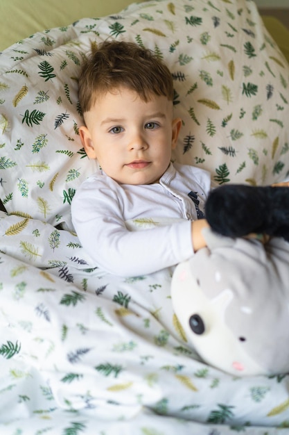 Niño pequeño feliz en pijama, en la cama en el dormitorio con juguetes, juegos, duerme. Hermosa ropa de cama para bebés. El sueño diurno de los niños. Luz tenue.