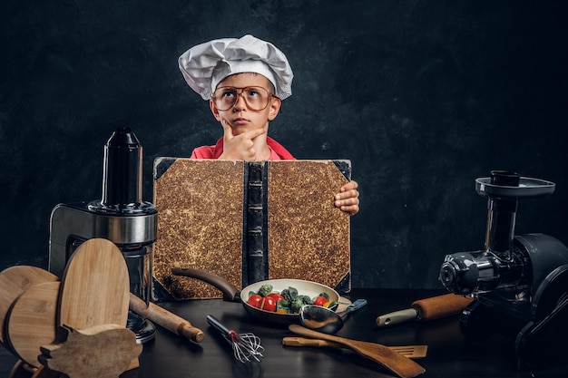 Un niño pequeño con gafas y sombrero de chef está leyendo un libro de recetas y planeando cocinar.