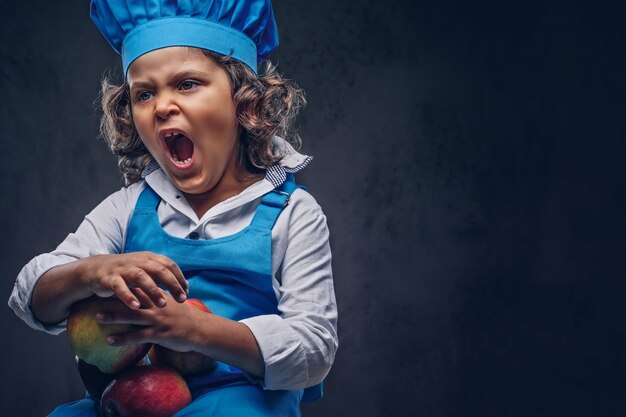 Un niño pequeño insatisfecho con el pelo rizado castaño vestido con un uniforme de cocinero azul sostiene manzanas en un estudio. Aislado en un fondo de textura oscura.