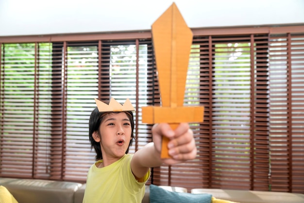 Un niño pequeño juega a un caballero sosteniendo una espada hecha con un pretexto de cartón para pelear en el período medieval con entusiasmo y alegría en la sala de estar en casa.