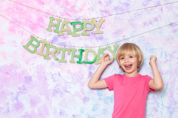 Niño pequeño lindo en camiseta rosa que adorna la pared colorida con palabras del feliz cumpleaños