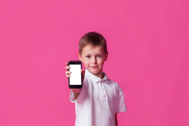 Niño pequeño lindo que muestra el teléfono celular de pantalla en blanco sobre fondo rosa