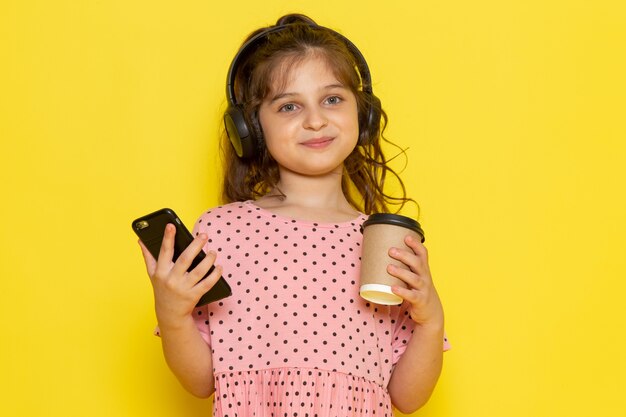 Un niño pequeño lindo de la vista frontal en vestido rosado que sostiene el teléfono inteligente y la taza de café