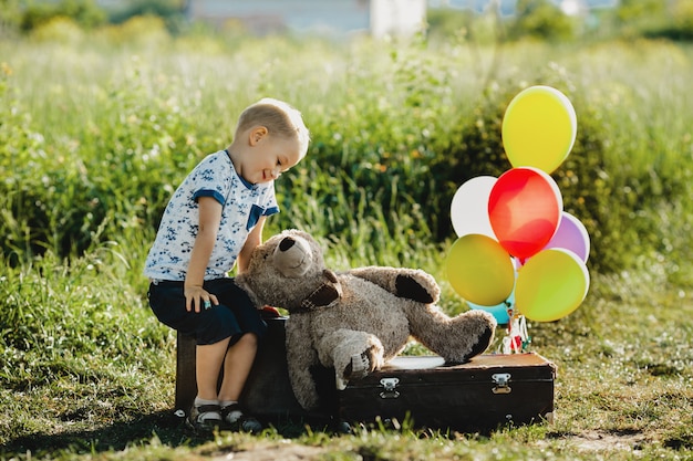 El niño pequeño con el oso de peluche se sienta en una maleta con los globos coloridos en el campo