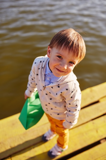 Niño pequeño que juega con el barco de papel del juguete por el lago