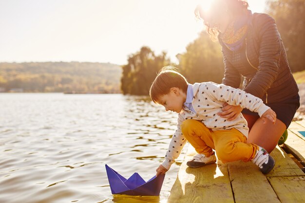 Niño pequeño que juega con el barco de papel del juguete por el lago