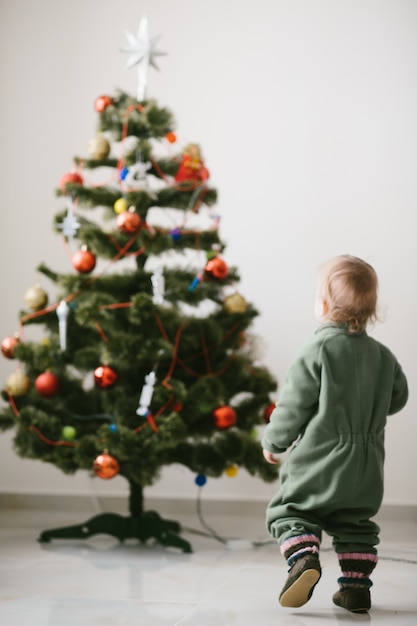 Niño pequeño en saltadores verdes camina al árbol de Navidad