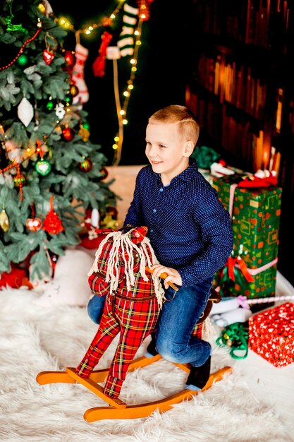 El niño pequeño se sienta en caballo rojo antes del árbol de Navidad